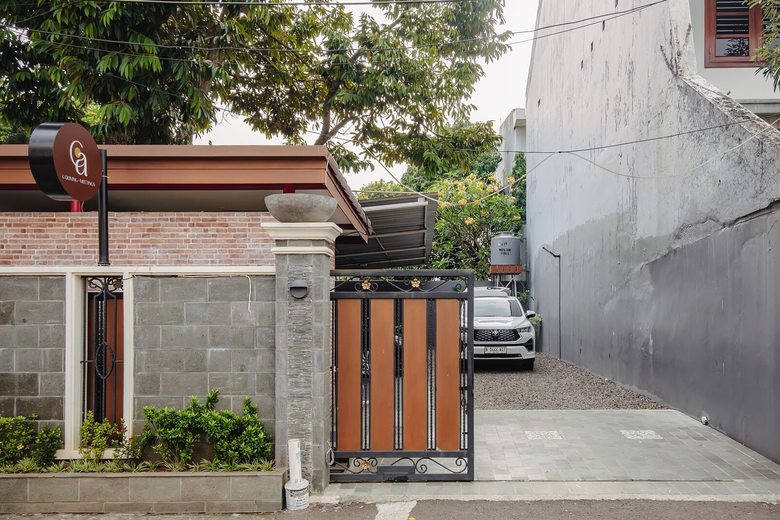 A gated entrance with a vehicle parked inside, a sign indicating a meeting point, and a building with a brick facade and a large tree in the background.