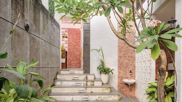 A narrow staircase with a concrete wall on one side and a brick wall on the other, leading to a door. There are potted plants and a hanging light fixture along the walls. - Calma Coliving at Bintaro, Tangerang Selatan (South Tangerang)