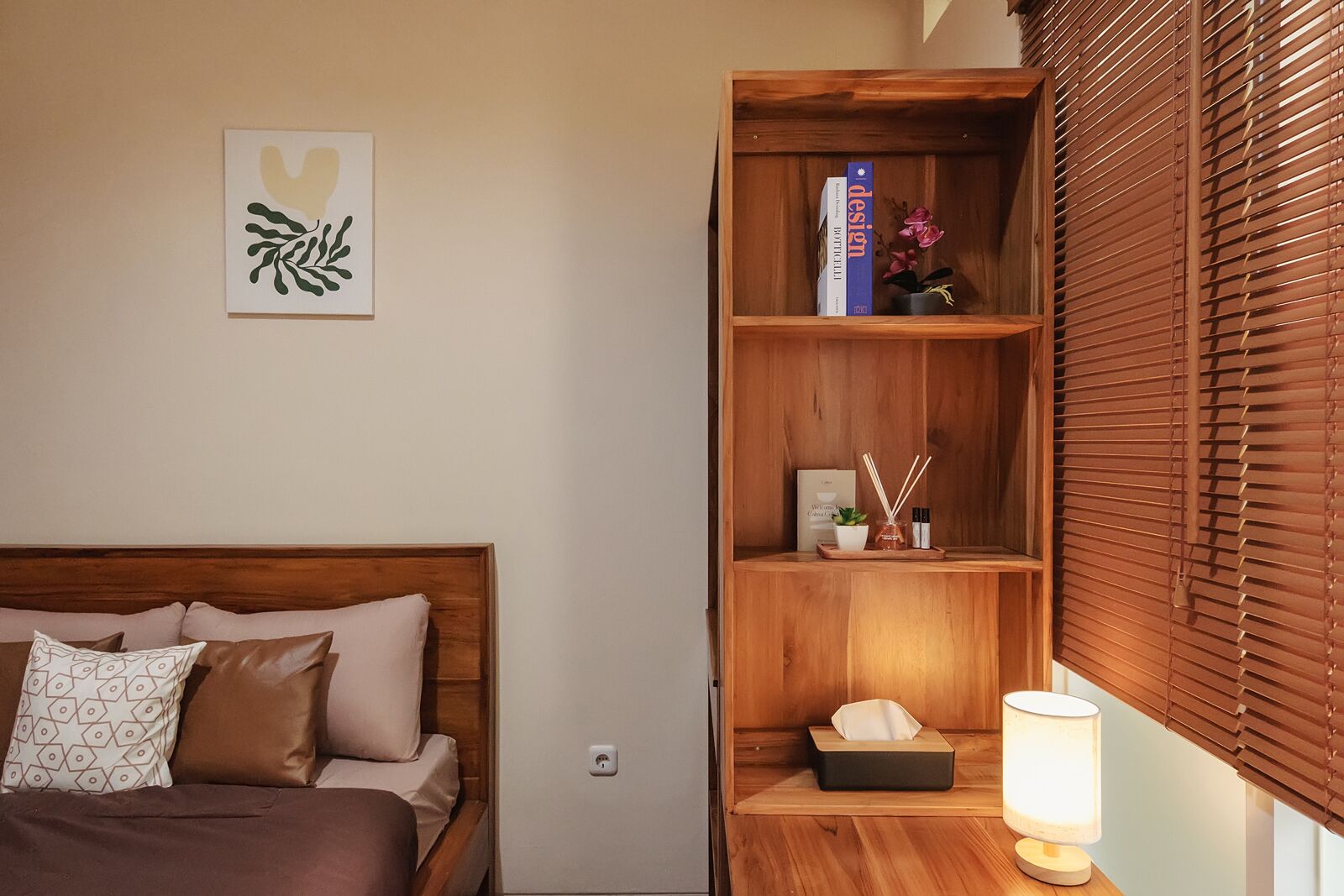 A bedroom with a wooden headboard, a bedside table with a lamp, a bookshelf, and a window with blinds.