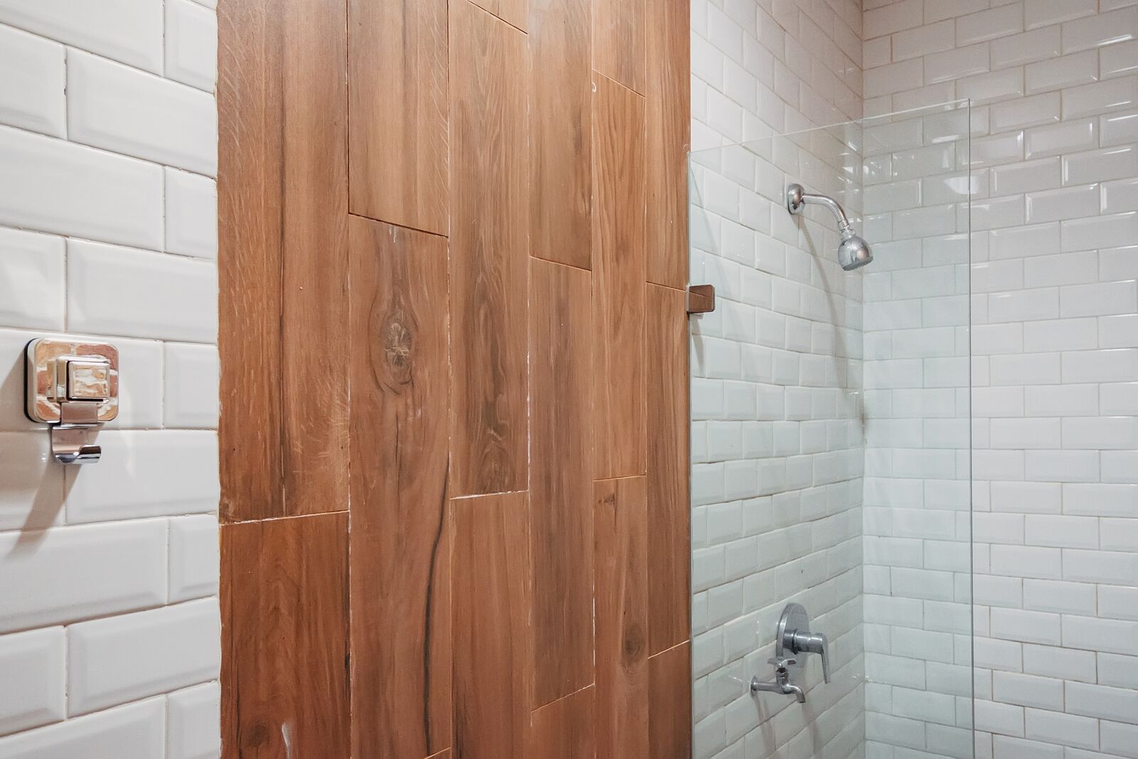 Une salle de bain avec des carreaux de céramique blancs rectangulaires, une porte en bois et une douche dotée d’une paroi en verre.