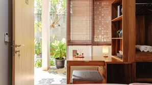 A room with a wooden desk, a bench, a bedside table, a wardrobe, and a view of a garden through a sliding door. - Calma Coliving at Bintaro, Tangerang Selatan (South Tangerang)