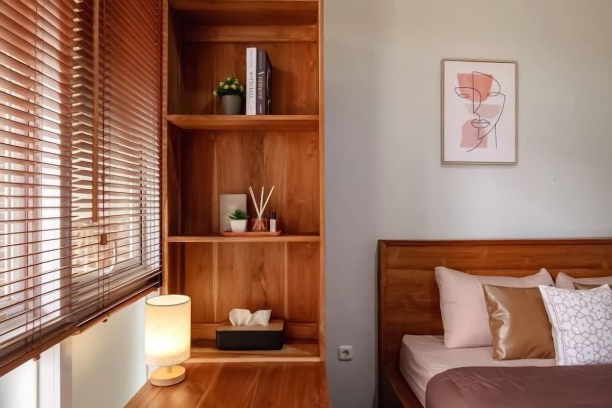 A bedroom with a wooden headboard, a bedside table with a lamp, a shelf with books and a small plant, and a framed artwork on the wall.