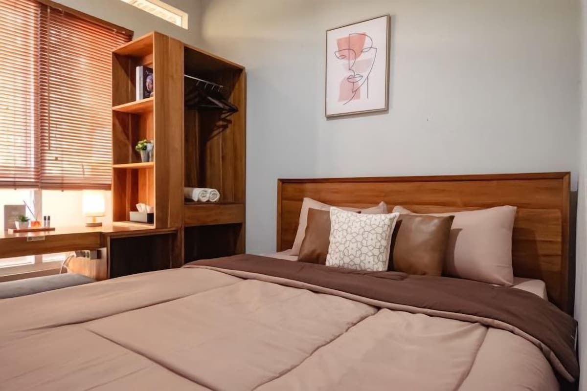 A bedroom with a wooden headboard, a bed with a brown blanket, a wooden wardrobe, and a framed artwork on the wall.