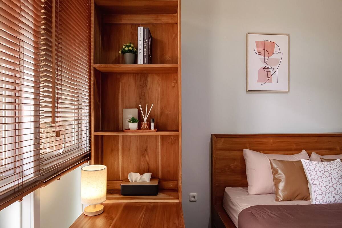 A bedroom with a wooden headboard, a bedside table with a lamp, a shelf with books and a plant, and a framed artwork on the wall.