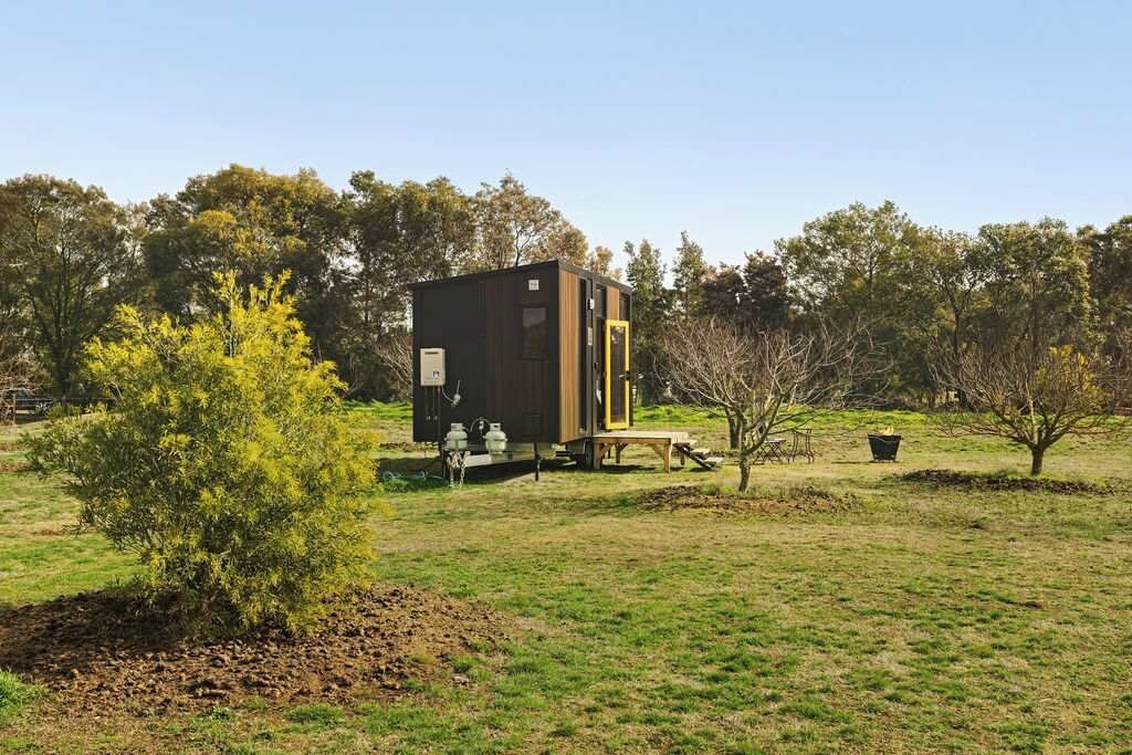 A small, dark-colored cabin with a wooden deck, surrounded by a grassy field and trees.