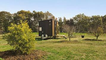 A small, dark-colored cabin with a wooden deck, surrounded by a grassy field and trees.