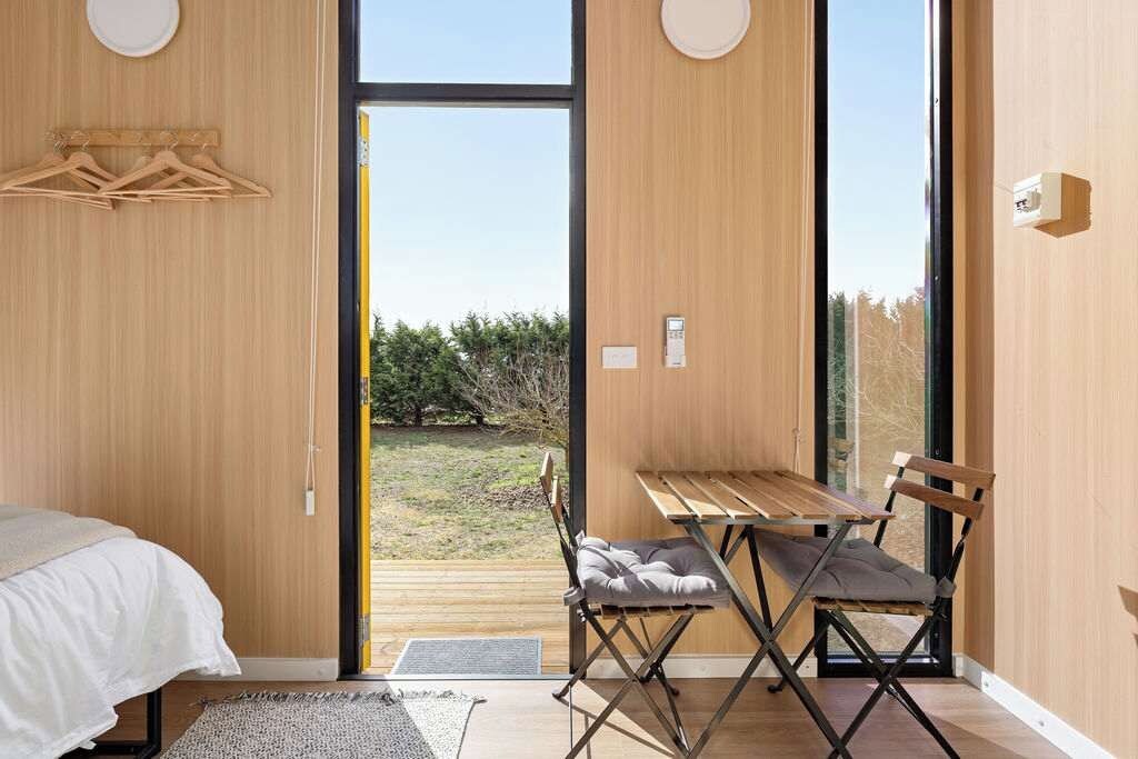 Un dormitori amb un llit, una taula de fusta amb una cadira, i vista a un jardí a través d'una porta de vidre.