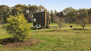 Eine kleine, dunkel gefärbte Holzhütte mit einer Holzterrasse, umgeben von einem grasbewachsen Feld und Bäumen.