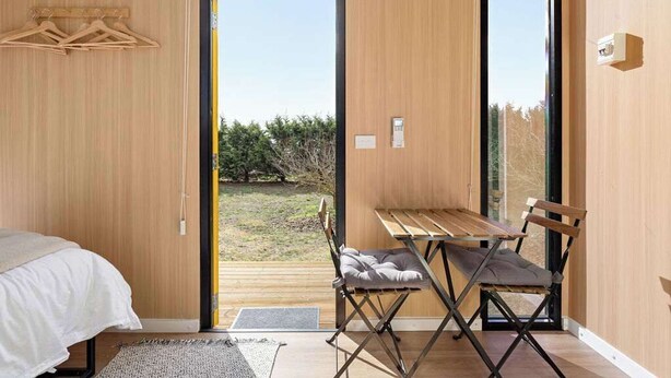 Ein Schlafzimmer mit einem Bett, einem Holztisch mit Stuhl und Blick auf einen Garten durch eine Glastür.