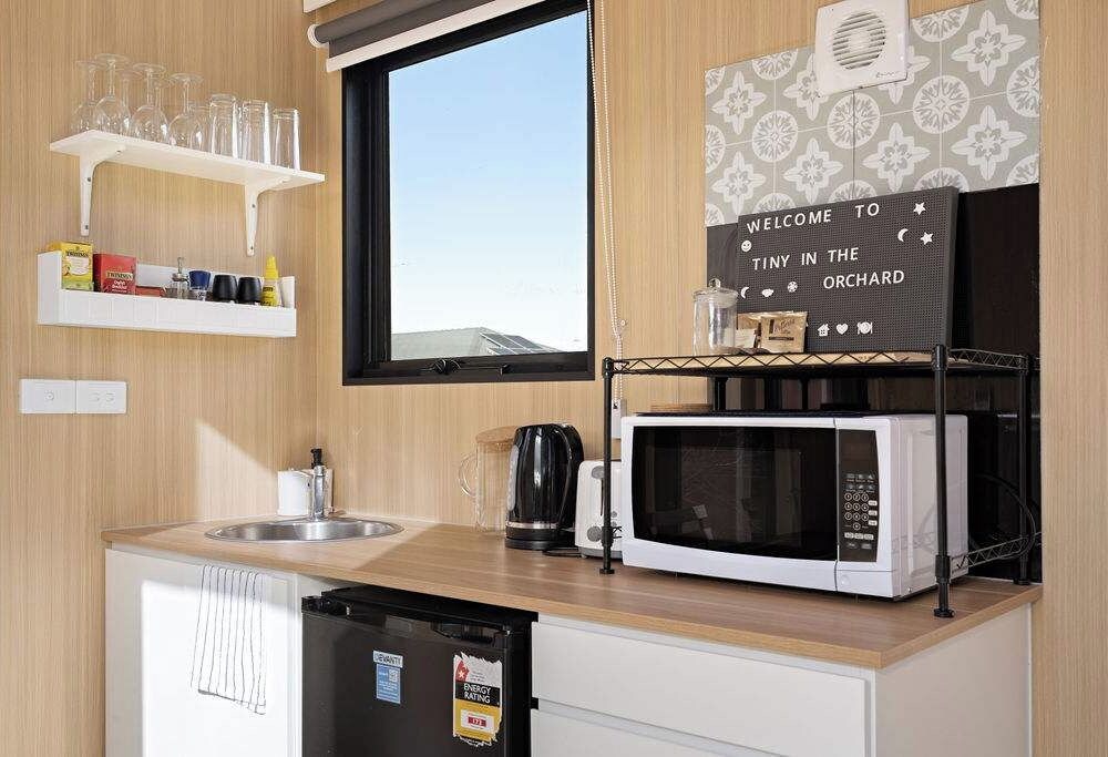 A compact kitchen with a microwave, kettle, and a shelf with glasses and condiments.