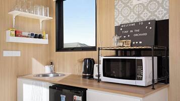 A compact kitchen with a microwave, kettle, and a shelf with glasses and condiments.