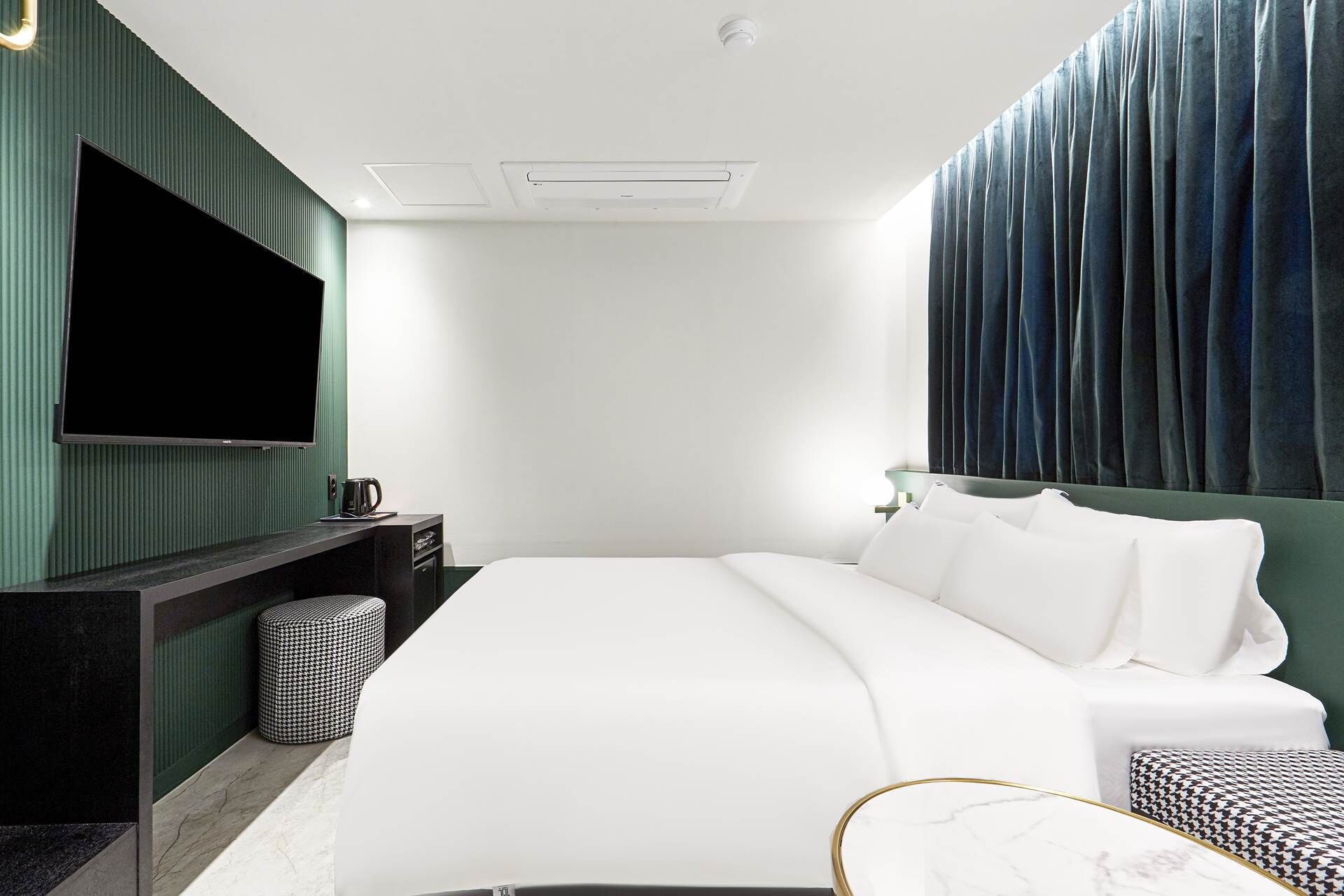 A modern hotel room with a large bed, a flat-screen TV, a small desk with a coffee maker, and a green accent wall.