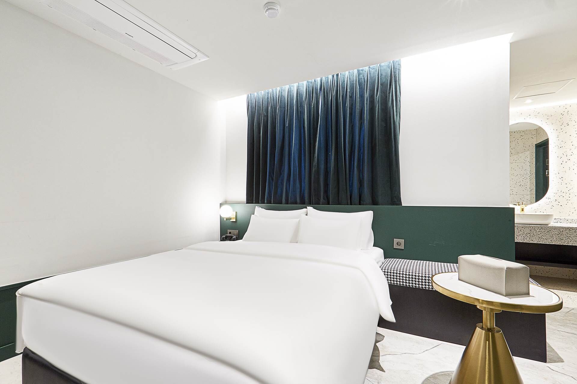Une chambre d’hôtel comprenant un lit, une table de chevet, une commode, une fenêtre avec des rideaux et une salle de bain avec un miroir.