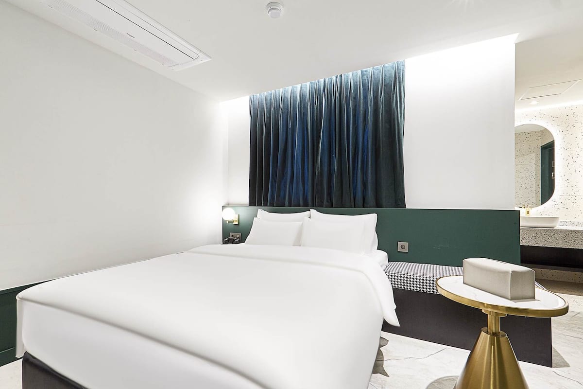 A hotel room with a bed, a bedside table, a nightstand, a window with curtains, and a bathroom with a mirror.