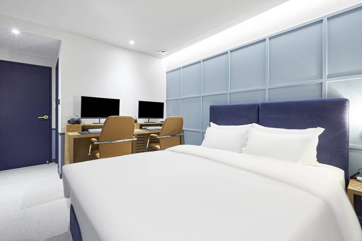 A hotel room with a bed, two desks with computers, and a large window.