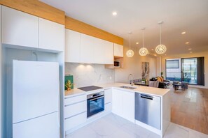 Una cocina moderna con armarios blancos, horno empotrado y lavavajillas. Hay luces colgantes sobre la isla y una zona de comedor con sofá y mesa.