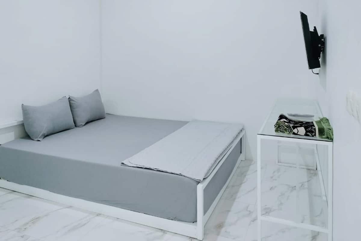 Quarto minimalista com cama cinza, paredes brancas, mesinha de cabeceira pequena com tecido estampado e televisão na parede.
