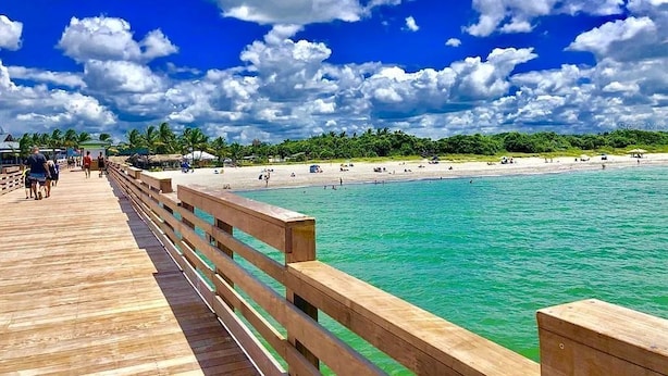 Eine Holzstege führt zu einem Strand mit klarem türkisblauem Wasser und einem blauem Himmel mit Wolken.
