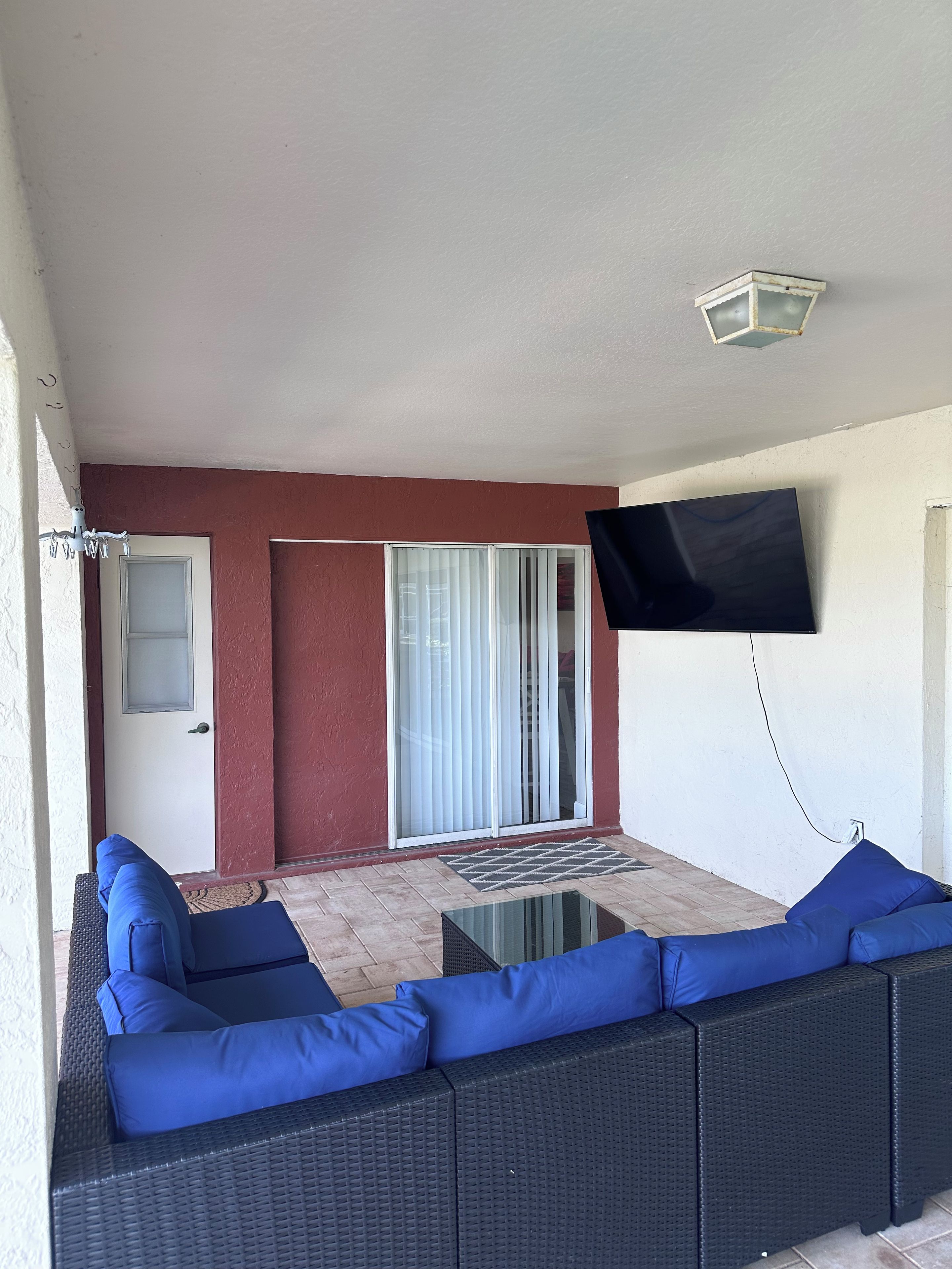Una habitación con un sofá de mimbre negro, un televisor montado en la pared y una puerta corrediza de vidrio.