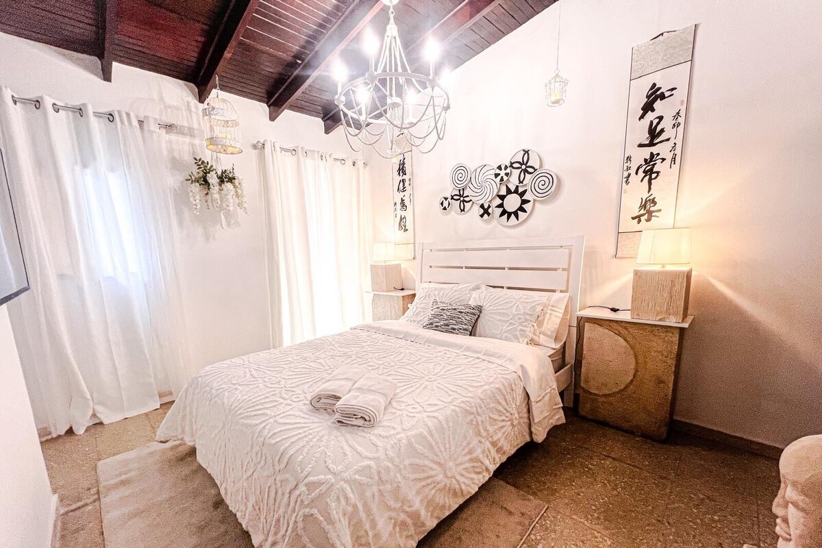 A bedroom with a large bed, a chandelier, a wall with calligraphy, and a small table with a lamp.