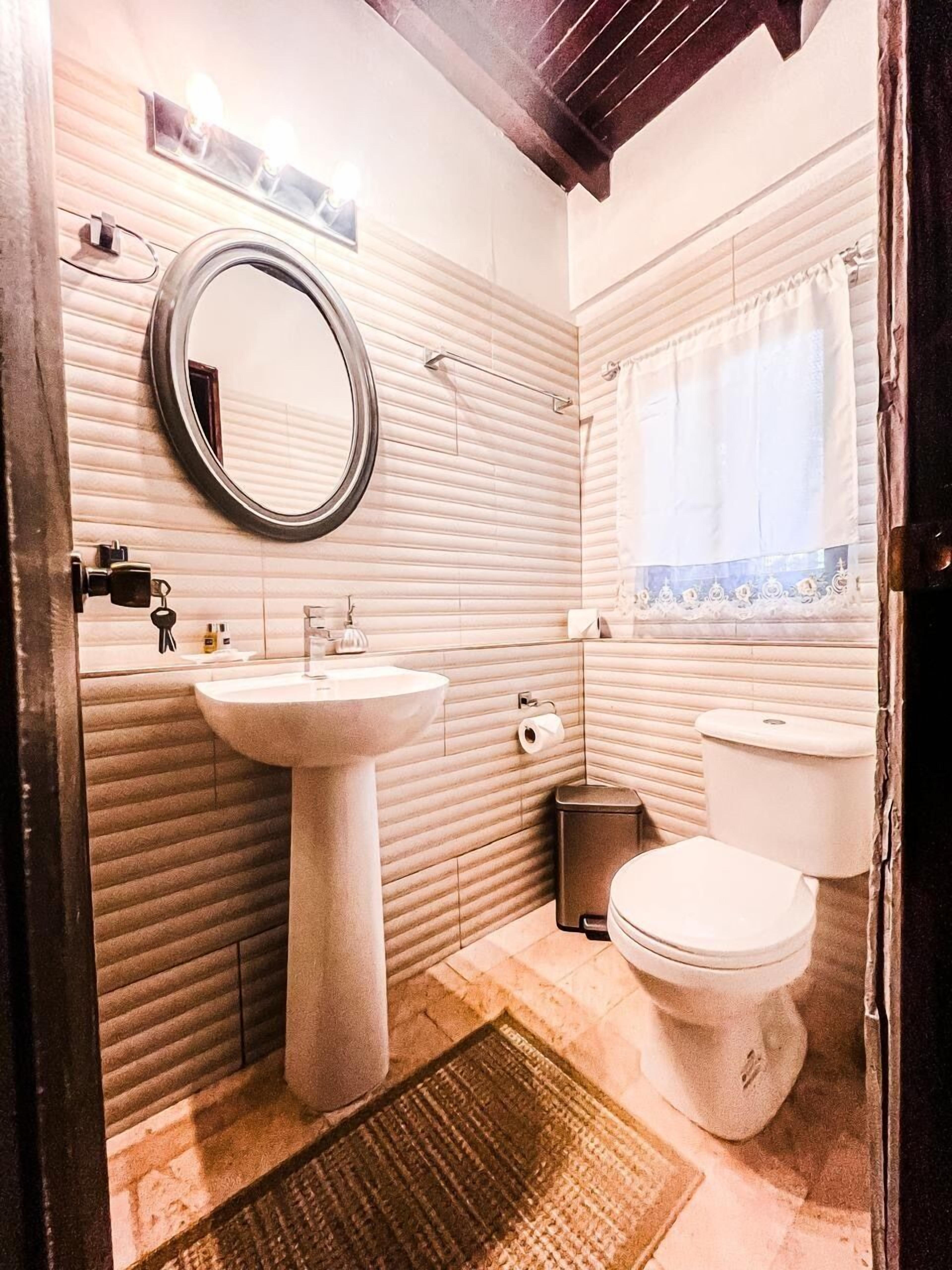 A bathroom with a pedestal sink, a round mirror, a toilet, and a window with curtains.