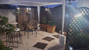 An outdoor patio area with a round table and two wicker chairs, surrounded by potted plants and a decorative wall.