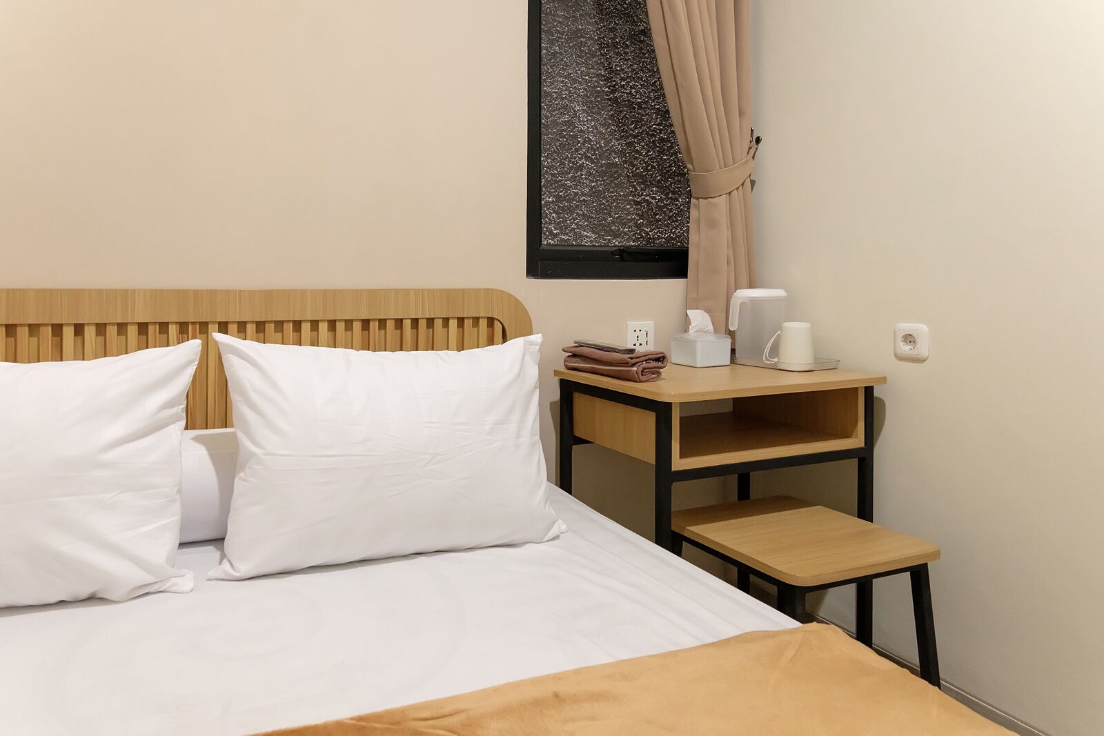 Cama bem arrumada com lençóis brancos e cabeceira de madeira, uma pequena mesa de cabeceira de madeira com caixa de lenços e cafeteira, e uma janela com cortina.