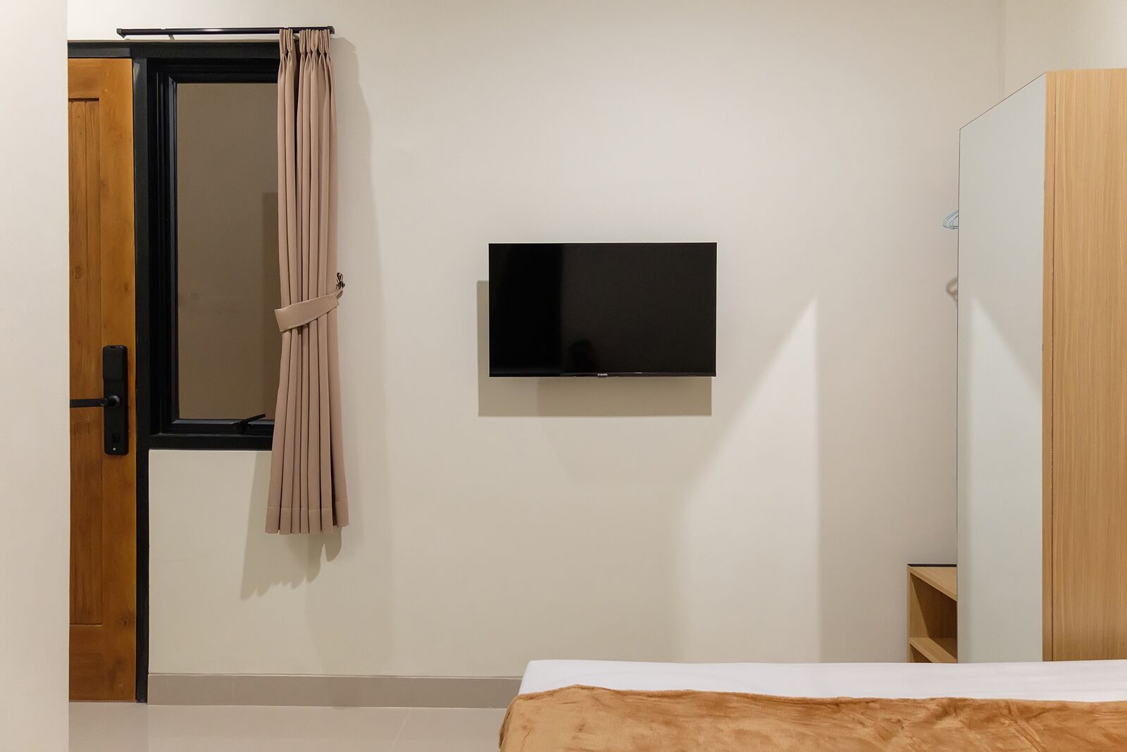 A bedroom with a bed, a TV mounted on the wall, a wooden wardrobe, and a door with a curtain.
