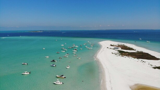 An aerial view of a beach with numerous boats anchored in turquoise waters, a sandy shoreline, and a clear blue sky.