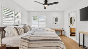 Une chambre à coucher comprenant un lit, un ventilateur de plafond, une table de chevet, une commode, une fenêtre avec des stores, une porte, une petite table avec une chaise et un téléviseur fixé au mur.