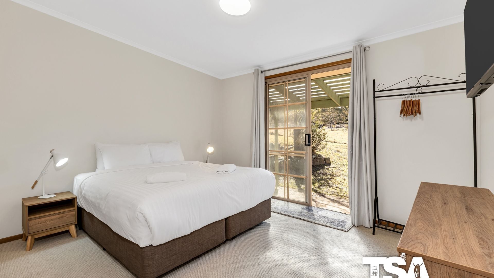 A bedroom with a large bed, a bedside table with a lamp, a wooden desk, and a view of the outdoors through a sliding glass door.
