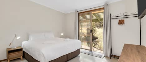 A bedroom with a large bed, a bedside table with a lamp, a wooden desk, and a view of the outdoors through a sliding glass door.