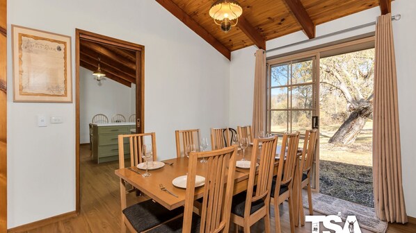Zona pranzo con tavolo e sedie in legno, un lampadario e vista sull'esterno attraverso un'ampia finestra.