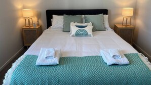A neatly made bed with a white comforter and two folded towels on a green patterned bedspread. There are two bedside tables with lamps, a dark headboard, and several pillows. - Cityscape minutes walk from all the city offers (Christchurch)