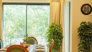 A dining area with a table set for four, surrounded by wooden chairs with green cushions. There are potted plants on the table and a large window with curtains.