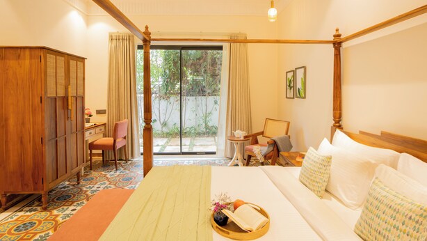A hotel room with a large bed, a wooden wardrobe, a desk with a chair, a small table, and a view of the outdoors through a sliding door.