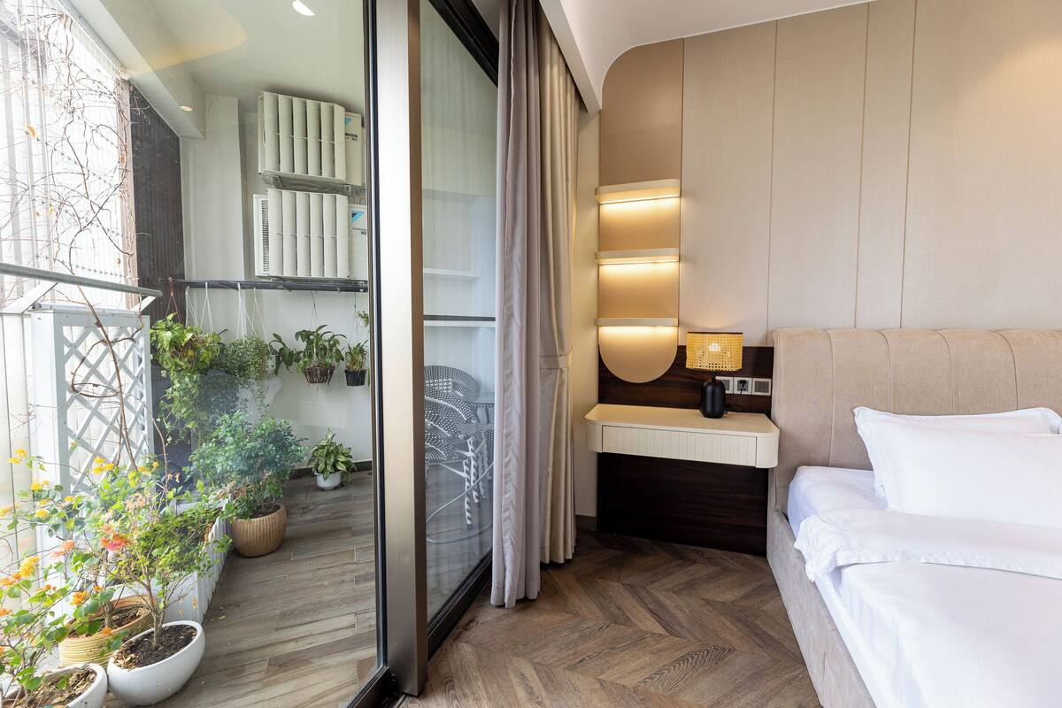 A modern hotel room with a bed, a small desk, a window with curtains, and potted plants.
