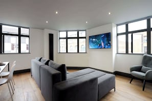A modern living room with a sectional sofa, a dining table with chairs, a TV mounted on the wall, and large windows.