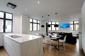 A modern kitchen with a central island, dining area with chairs, a black sofa, and a TV mounted on the wall.