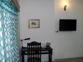 Um quarto com uma mesa de madeira, uma cadeira, um quadro emoldurado, uma televisão fixada na parede e um arandela.