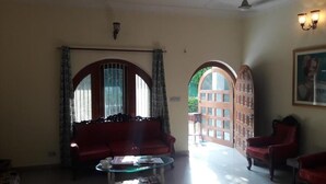 Una sala de estar con un sofá rojo, una mesa de centro de vidrio y un sillón rojo. Hay dos ventanas arqueadas grandes con cortinas, y una puerta que da al exterior.