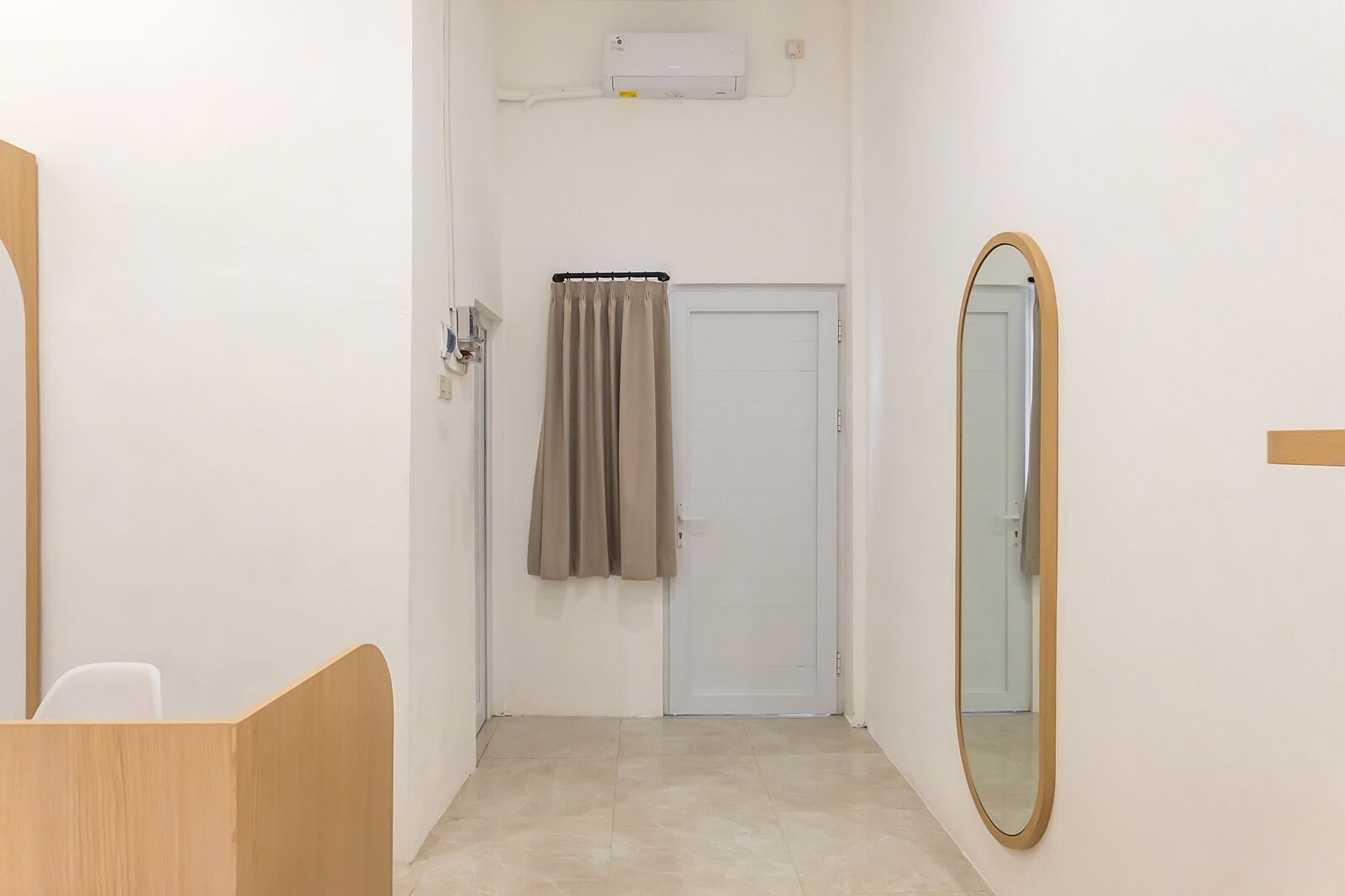 A narrow hallway with a white door, a towel hanging on the door, a mirror on the right, and a small white chair in the foreground.