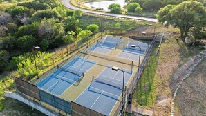 An outdoor tennis court with a surrounding fence, a river in the background, and a road curving away. - Relaxing Lake Escape | 1BR | Pickleball | BBQ (Lake Travis)