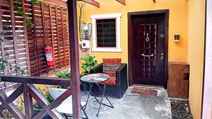Un patio con una pérgola de madera, un extintor, una mesa pequeña con una silla, y una puerta con cerradura electrónica.