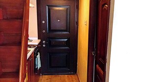 Un estrecho pasillo de madera con una puerta oscura al final, una pequeña zona de cocina con un lavamanos y armarios, y un suelo de madera.