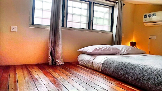 Un dormitorio con piso de madera, cama con ropa blanca, ventana con cortinas y un equipo de aire acondicionado en la pared.
