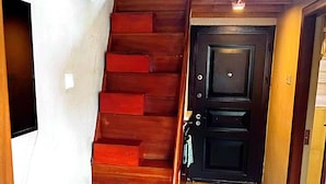 Una escalera de madera que conduce a una puerta oscura, con un interruptor de luz en la pared y un ventanal arriba.