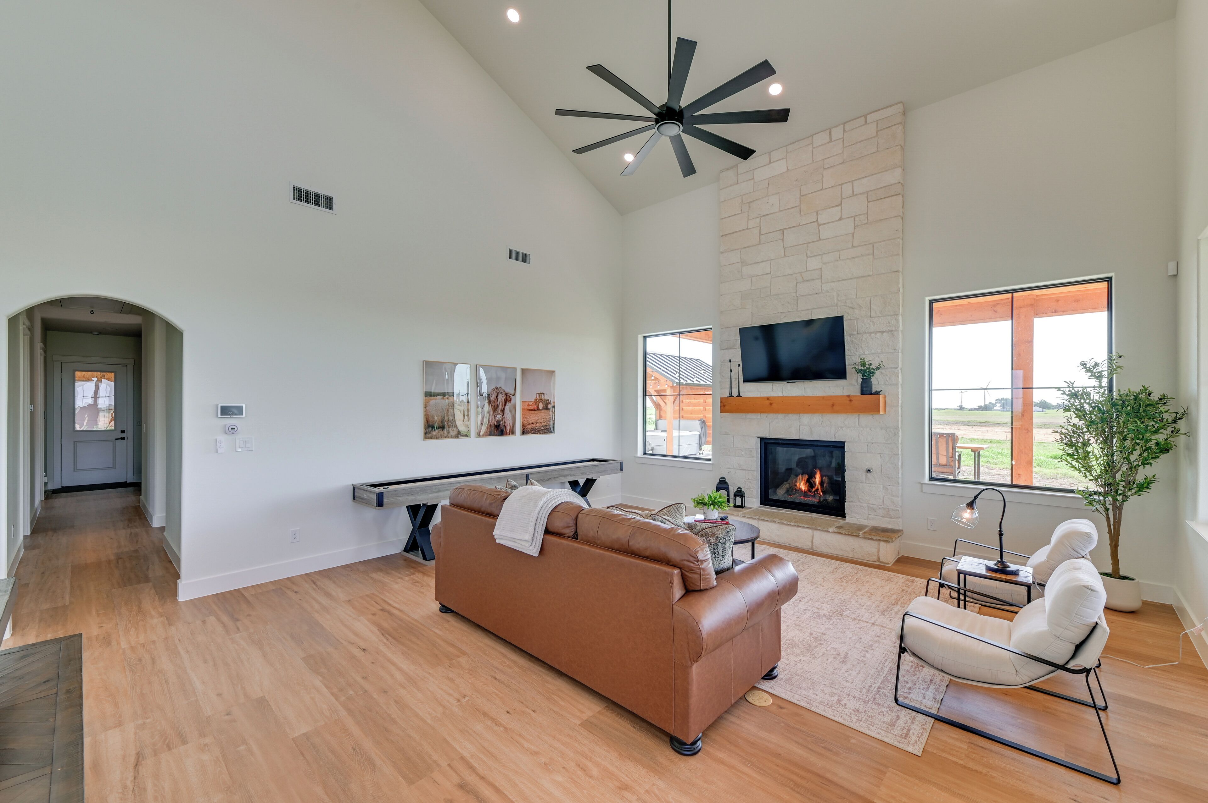 A spacious living room with a brown leather sofa, a white armchair, a fireplace, a TV mounted on a brick wall, and a large window offering a view of the outdoors.