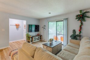A spacious living room with a large sofa, a coffee table, and a television. There is a sliding glass door leading to an outdoor area with plants.