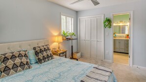 Un dormitorio con cama, ventilador de techo, mesita de noche con lámpara, ventana con persianas y puerta que da a un baño.